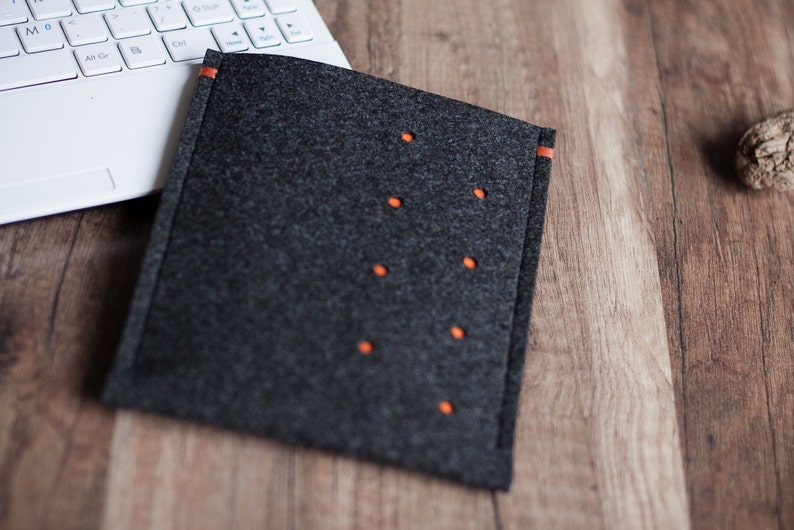 May include: A gray felt laptop sleeve with orange stitching and decorative stitching. The sleeve is lying on a wooden surface.