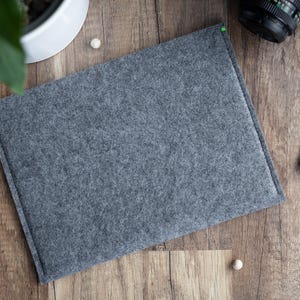May include: A rectangular, dark gray felt sleeve with a textured surface and stitched edges. The sleeve features small, bright green accent threads at the top corners, providing a subtle contrast to the neutral gray material of the flat pouch.