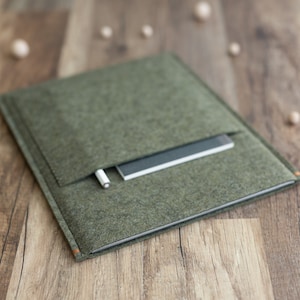 May include: A green felt laptop sleeve with a pocket for a pen or pencil. The sleeve is lying on a wooden surface.