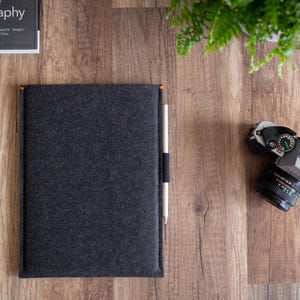 Peut inclure: Une pochette en feutre gris fonc&eacute; pour tablette ou ordinateur portable, dot&eacute;e d'un stylet blanc vertical maintenu par une boucle &eacute;lastique noire sur le c&ocirc;t&eacute; droit, avec de petits accents orange aux coins sup&eacute;rieurs, pos&eacute;e sur une surface en bois.