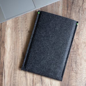 May include: A dark gray felt laptop sleeve with green accents at the top corners. The sleeve is rectangular and lies on a wooden surface next to a laptop.