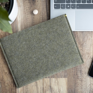 Könnte beinhalten: Eine grüne Filz-Laptoptasche mit gelben Nähten. Die Tasche liegt auf einer Holzoberfläche neben einem Laptop, einem Handy und einer Topfpflanze.