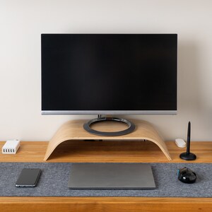 Op de afbeelding: Een monitorstandaard van licht hout met een gebogen ontwerp staat op een houten bureau. Een computermonitor, een grijze laptop, een zwarte muis, een smartphone, een stylus op een standaard en een grote grijze vilten bureaumat zijn aanwezig.