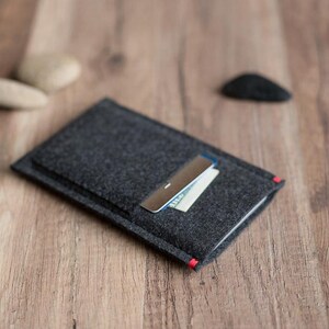 May include: A dark gray felt wallet with a silver money clip holding a white card. The wallet has a red accent on the corner. The wallet is on a wooden surface.