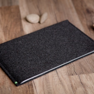 May include: A dark gray felt laptop sleeve with green stitching. The sleeve is lying on a wooden surface.