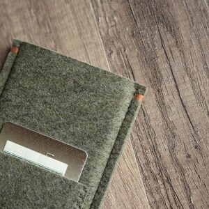 May include: An olive green felt card holder featuring a rectangular pocket with visible stitching and orange thread accents at the top corners. A metallic gold credit card with a geometric pattern is partially inserted into the front pocket.