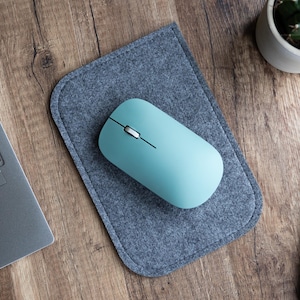 May include: A light blue wireless computer mouse on a gray felt mousepad. The mouse is on a wooden desk with a laptop and a digital clock.