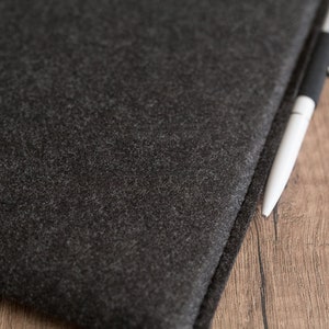 Peut inclure: Une pochette en feutre gris anthracite fonc&eacute; avec une surface &eacute;paisse et textur&eacute;e aux bords cousus. Un stylo &agrave; bille &eacute;l&eacute;gant avec un corps blanc et une prise noire est plac&eacute; le long du bord de la pochette en feutre, sur une surface en bois.