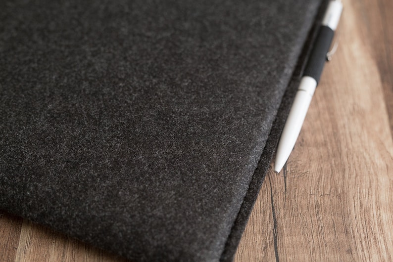 Peut inclure: Une pochette en feutre gris anthracite fonc&eacute; avec une surface &eacute;paisse et textur&eacute;e aux bords cousus. Un stylo &agrave; bille &eacute;l&eacute;gant avec un corps blanc et une prise noire est plac&eacute; le long du bord de la pochette en feutre, sur une surface en bois.