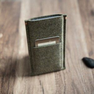 May include: A rectangular olive green felt sleeve designed to hold a mobile device, featuring a smaller exterior pocket on the front containing a single silver-colored rectangular card. The sleeve has visible stitching along the edges and sides.