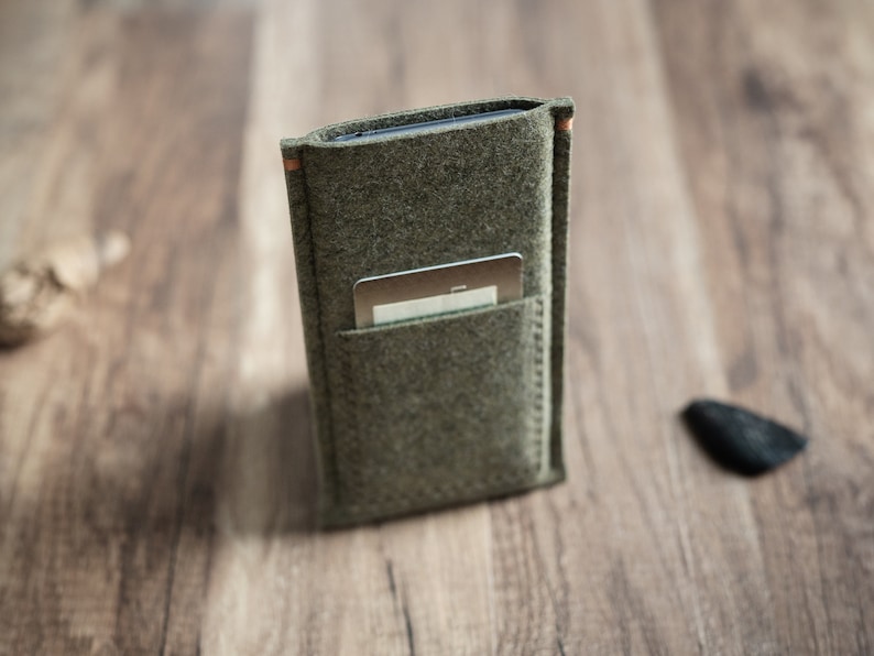 May include: A rectangular olive green felt sleeve designed to hold a mobile device, featuring a smaller exterior pocket on the front containing a single silver-colored rectangular card. The sleeve has visible stitching along the edges and sides.