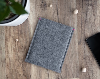 May include: A grey felt sleeve with a pink stitch detail, possibly for a tablet or e-reader, sits on a wooden surface. A smartphone and a potted plant are also visible. The sleeve's minimalist design suggests a focus on protection and style.