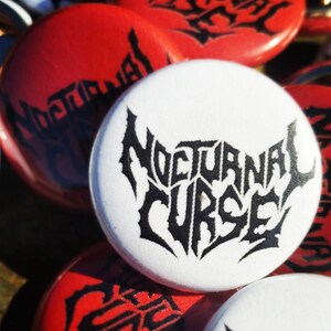 May include: A collection of round buttons with the words "Nocturnal Curse" in a gothic font. The buttons are white with black text and red with black text. The buttons are likely for a band or related merchandise.