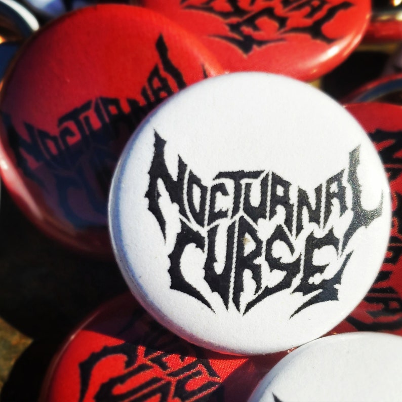 May include: A collection of round buttons with the words "Nocturnal Curse" in a gothic font. The buttons are white with black text and red with black text. The buttons are likely for a band or related merchandise.
