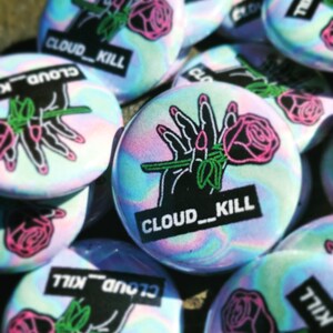 Op de afbeelding: Een verzameling iriserende holografische knopen met een zwart-witte afbeelding van een hand die een roze roos vasthoudt en de tekst "CLOUD_KILL".