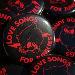 May include: Black button with red text and a panther graphic. The text reads "LOVE SONGS FOR JUNKIES" with a lightning bolt. The buttons are glossy and have a retro, punk rock aesthetic.