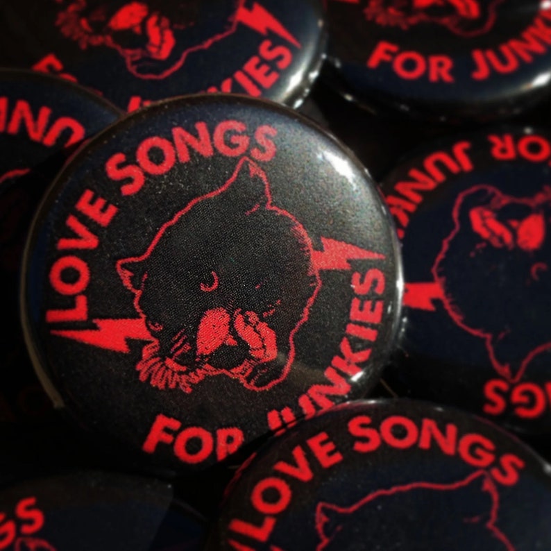 May include: Black button with red text and a panther graphic. The text reads "LOVE SONGS FOR JUNKIES" with a lightning bolt. The buttons are glossy and have a retro, punk rock aesthetic.