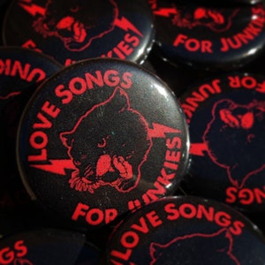 Op de afbeelding: Zwarte en rode knoopsbadge met een cartoonbeer en een bliksem. De tekst luidt "Love Songs For Junkies".