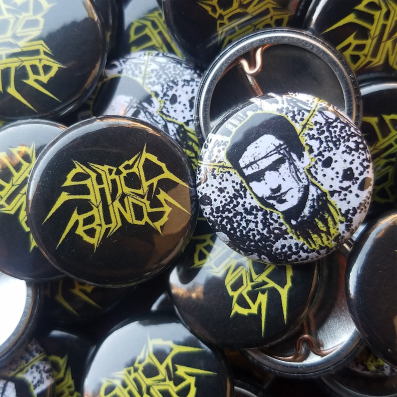 May include: A collection of black button pins with yellow and white graphics. Some pins feature the text "SHRED BUNNY" in a stylized font. Other pins display a black and white portrait. The pins are round.