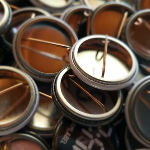 May include: A close-up shot of several metal pin-back buttons. The buttons have a shiny, metallic finish with a safety pin on the back. The buttons are in various shades of silver and gold, with some showing the back of the button.