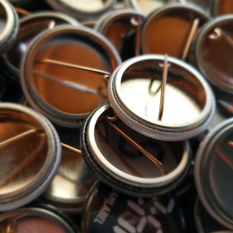 May include: A close-up shot of several metal pin-back buttons. The buttons have a shiny, metallic finish with a safety pin on the back. The buttons are in various shades of silver and gold, with some showing the back of the button.