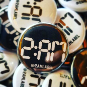May include: A collection of round buttons with a digital clock face design. The central button displays "2:01 AM" in white, with "@2AMLABEL" below. Other buttons show similar digital clock designs in white on black or white backgrounds.