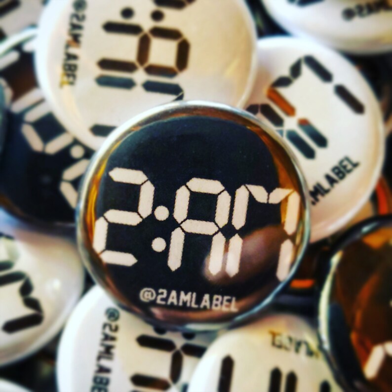 May include: A collection of round buttons with a digital clock face design. The central button displays "2:01 AM" in white, with "@2AMLABEL" below. Other buttons show similar digital clock designs in white on black or white backgrounds.