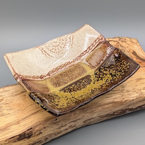 May include: A handmade ceramic rectangular serving dish with a unique design. The dish features a light beige section with a textured pattern, a brown section, and a dark brown section with gold speckles. The edges are slightly wavy.