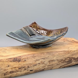 May include: A handmade ceramic bowl with a unique, asymmetrical design. The bowl features a dark blue-grey exterior with a textured pattern on the sides and a brown and gold speckled interior. The bowl is displayed on a piece of wood.