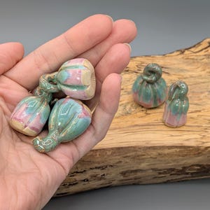 May include: A collection of ceramic sculptures in the shape of pumpkins and gourds. The pieces are glazed in shades of green, pink, and cream. The sculptures vary in size and shape, with some featuring twisted stems. The background is a wooden surface.
