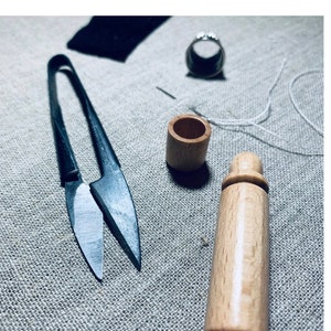 May include: A close-up of sewing tools on a textured, neutral-toned surface. The image features a pair of black scissors, a wooden thread holder, a wooden needle case, a ring, and a piece of black fabric. The text "Thimble & Plume" is at the bottom.