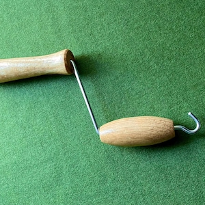 May include: A wooden handle with a metal hook, likely a tool for pulling or hanging items. The handle is light brown, and the hook is silver. The tool is set against a green background.