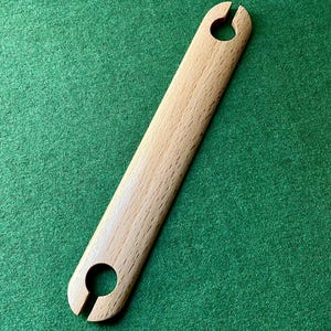 Pode incluir: Uma lançadeira de tecelagem de madeira clara, com formato retangular arredondado. Possui um orifício circular com uma fenda em cada extremidade. A lançadeira está sobre um fundo verde.