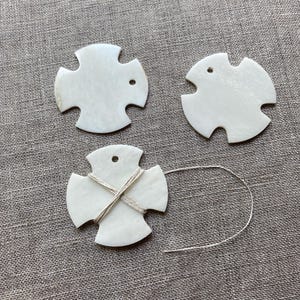 May include: Three off-white, cross-shaped pendants with rounded edges. One pendant has twine wrapped around it. Each pendant has a small hole at the top. The pendants are on a textured, gray fabric surface.
