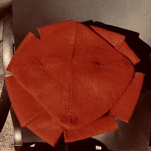 May include: A rust-colored felt hat with a unique, folded design. The hat is folded into eight sections, creating a geometric pattern.