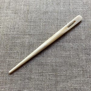 May include: A long, slender, off-white bone needle with a rectangular eyelet near the top. The needle tapers to a fine point. The item is likely used for sewing or crafting. The background is a textured, neutral-colored fabric.