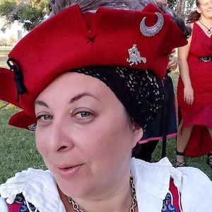 May include: A woman wearing a red hat with a silver crescent moon and a feather. The hat is decorated with a silver pin of a knight riding a horse. She is wearing a white shirt with a silver chain necklace and blue and silver buttons.