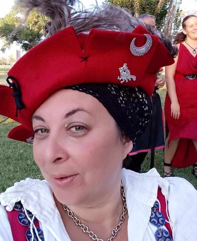 May include: A woman wearing a red hat with a silver crescent moon and a feather. The hat is decorated with a silver pin of a knight riding a horse. She is wearing a white shirt with a silver chain necklace and blue and silver buttons.
