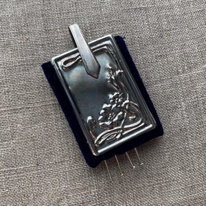 Op de afbeelding: Een zilverkleurig rechthoekig speldenkussen met een sierlijk bloemmotief en een handvat. Het speldenkussen staat op een donkerblauwe fluwelen basis, met verschillende spelden erin.