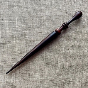 Könnte beinhalten: Ein dunkelbrauner Brieföffner mit rundem Griff und langer, spitz zulaufender Klinge. Der Griff hat ein bauchiges Ende und ein dekoratives, gedrehtes Design. Der Brieföffner ist aus Holz gefertigt und liegt auf einer strukturierten, neutralfarbenen Oberfläche.