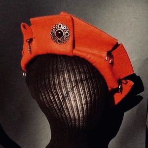 May include: A red felt hat with a decorative gold and red jewel embellishment. The hat is worn on a black mannequin head.