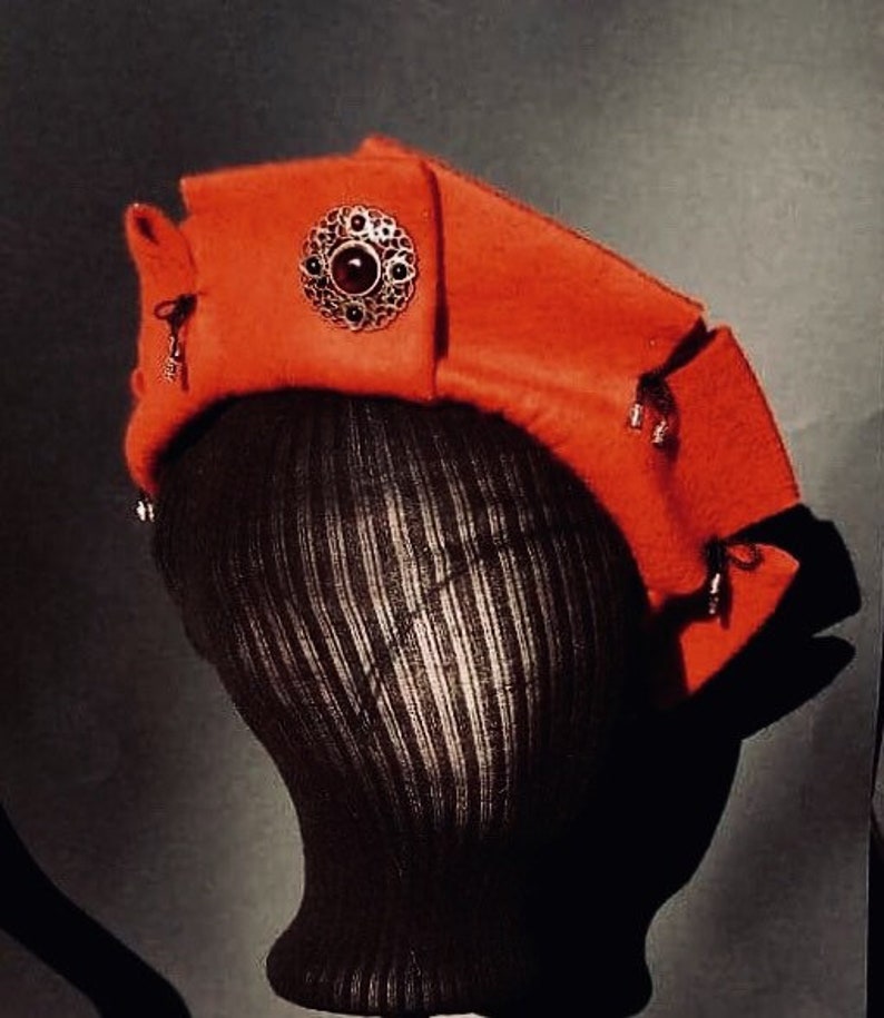 May include: A red felt hat with a decorative gold and red jewel embellishment. The hat is worn on a black mannequin head.