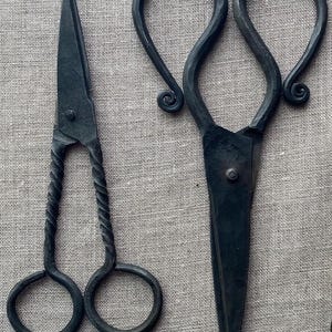 Puede incluir: Dos pares de tijeras de metal negro. Un par tiene un mango retorcido y agujeros redondos para los dedos. El otro par tiene un mango en forma de lazo con remolinos decorativos. Ambos tienen hojas puntiagudas.