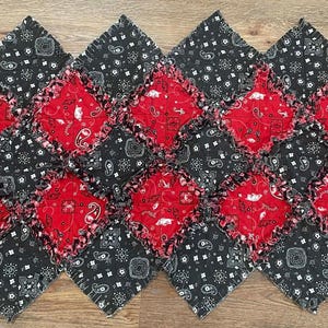 May include: A handmade, patchwork table runner featuring a repeating pattern of red and black bandana fabric squares. The red squares are centered within the black squares, creating a visually appealing design. The edges are frayed for a rustic look.