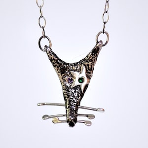 Puede incluir: Un collar de plata con un colgante texturizado en forma de cara de animal estilizada con piedras preciosas verdes y moradas para los ojos.