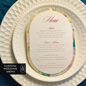 May include: A custom wedding menu card with an oval shape, featuring a floral design and elegant script. The menu is placed on a white plate with a textured rim. The menu lists starters and dinner stations.
