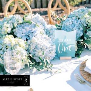 Puede incluir: Decoración de mesa de boda con hortensias azul claro. Se muestra un número de mesa de acrílico transparente con las palabras "TABLE One". También se ven una copa de vino y un mantel individual tejido.