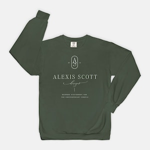 May include: Dark green sweatshirt with the text "ALEXIS SCOTT designs" in white script. Below the text is the phrase "BESPOKE STATIONERY FOR THE CONTEMPORARY COUPLE". The sweatshirt has long sleeves and a crew neck.