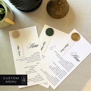 May include: Three white cards with black calligraphy text, each with a wax seal in a different color: gold, green, and beige. The text on the cards reads "Menu" and lists a wedding salad, starter, entree, and dessert. The text also includes the words "Custom Menu".