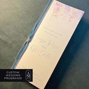 May include: A custom wedding program with a light pink background and a watercolor floral design in shades of purple and yellow. The program is tied with a light blue ribbon. The text reads "Welcome to the Wedding of Surra Price and Kyle Carter."